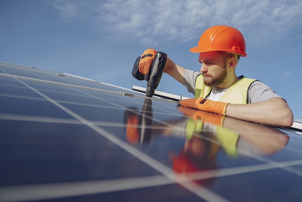 Low angle of serious bearded male electrician in hardhat and protective gloves installing solar photovoltaic panel system using drill